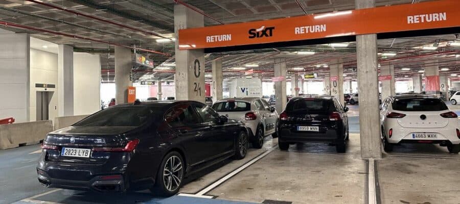 Sixt rental car return area in a covered parking garage, featuring a black BMW sedan, SUVs, and compact cars parked under an orange Return sign.